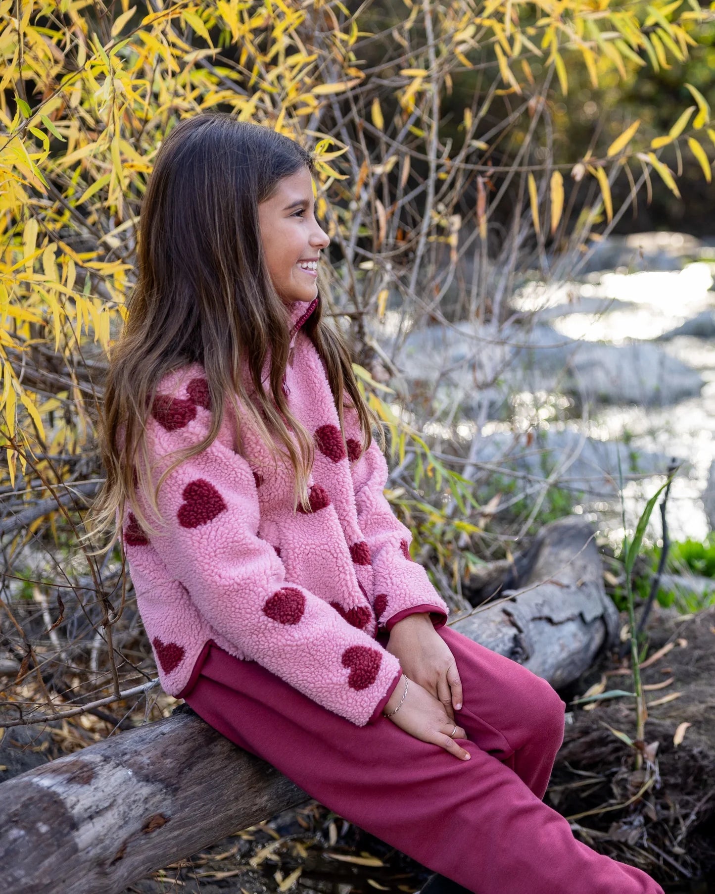 A young girl sitting outdoors on a log by a stream, wearing a pink fleece jacket with dark red heart patterns and matching pink pants.