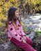 A young girl sitting outdoors on a log by a stream, wearing a pink fleece jacket with dark red heart patterns and matching pink pants.