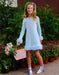A young girl stands outdoors in front of a garden of potted plants, wearing a light blue long-sleeve tunic dress with fluffy trim at the cuffs and hem. She is holding a pink handbag with a rainbow cloud patch and wearing shiny silver boots with purple socks.