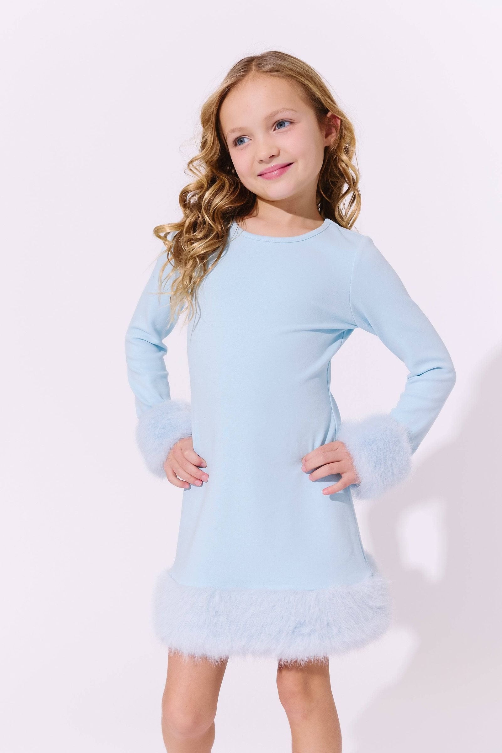 A young girl with long wavy blonde hair wearing a light blue long-sleeve dress with fuzzy trim on the cuffs and hem, posing against a plain white studio background.