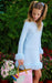 A young girl with blonde pigtails stands in front of a lush green garden background, wearing a light blue long-sleeve dress with fluffy trim and holding a pink handbag with gold handles and a rainbow cloud patch.