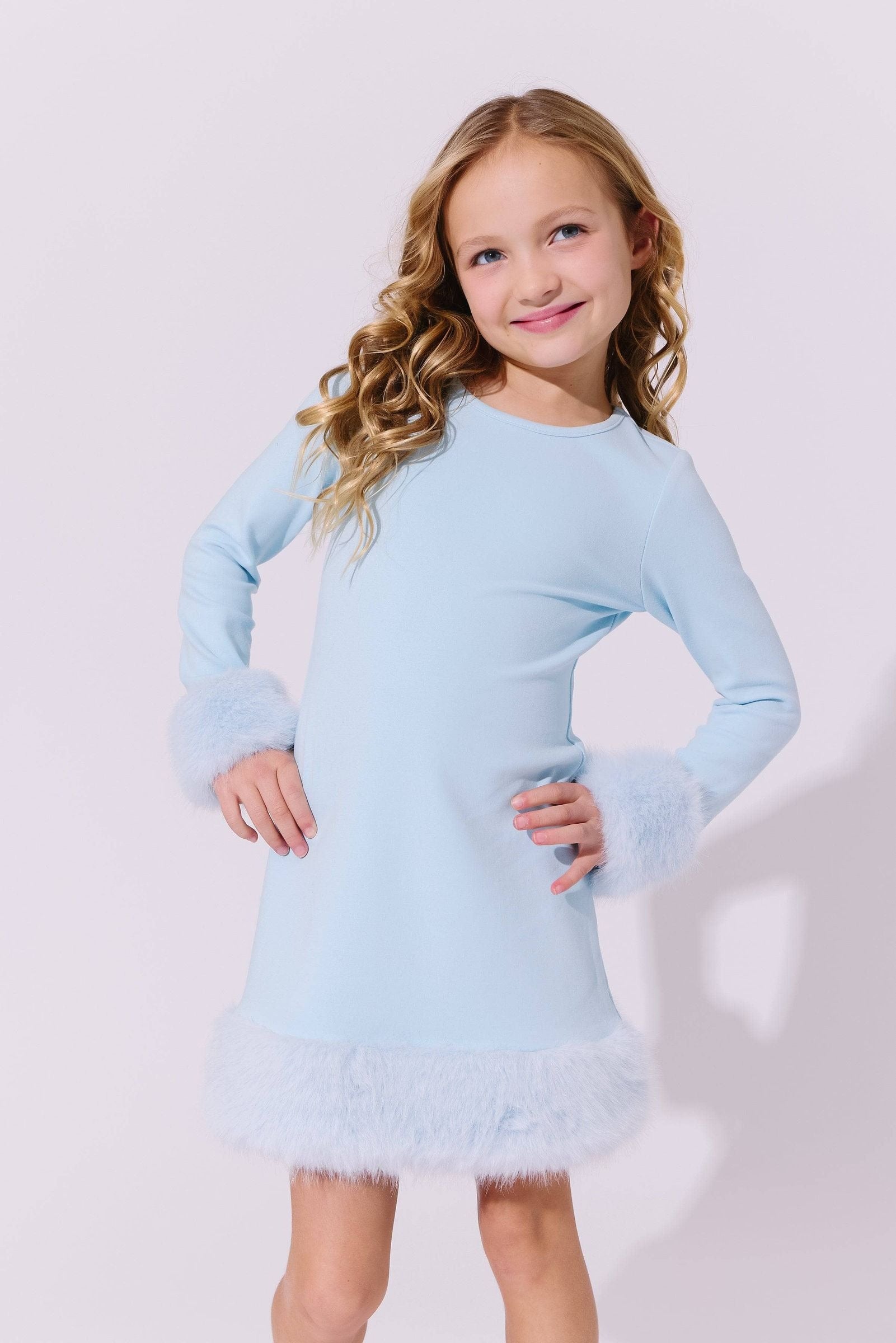 A young girl with blonde curly hair wearing a light blue long-sleeve dress with fluffy faux-fur trim at the cuffs and hem, posing against a light gray studio background.