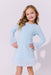 A young girl with blonde curly hair wearing a light blue long-sleeve dress with fluffy faux-fur trim at the cuffs and hem, posing against a light gray studio background.