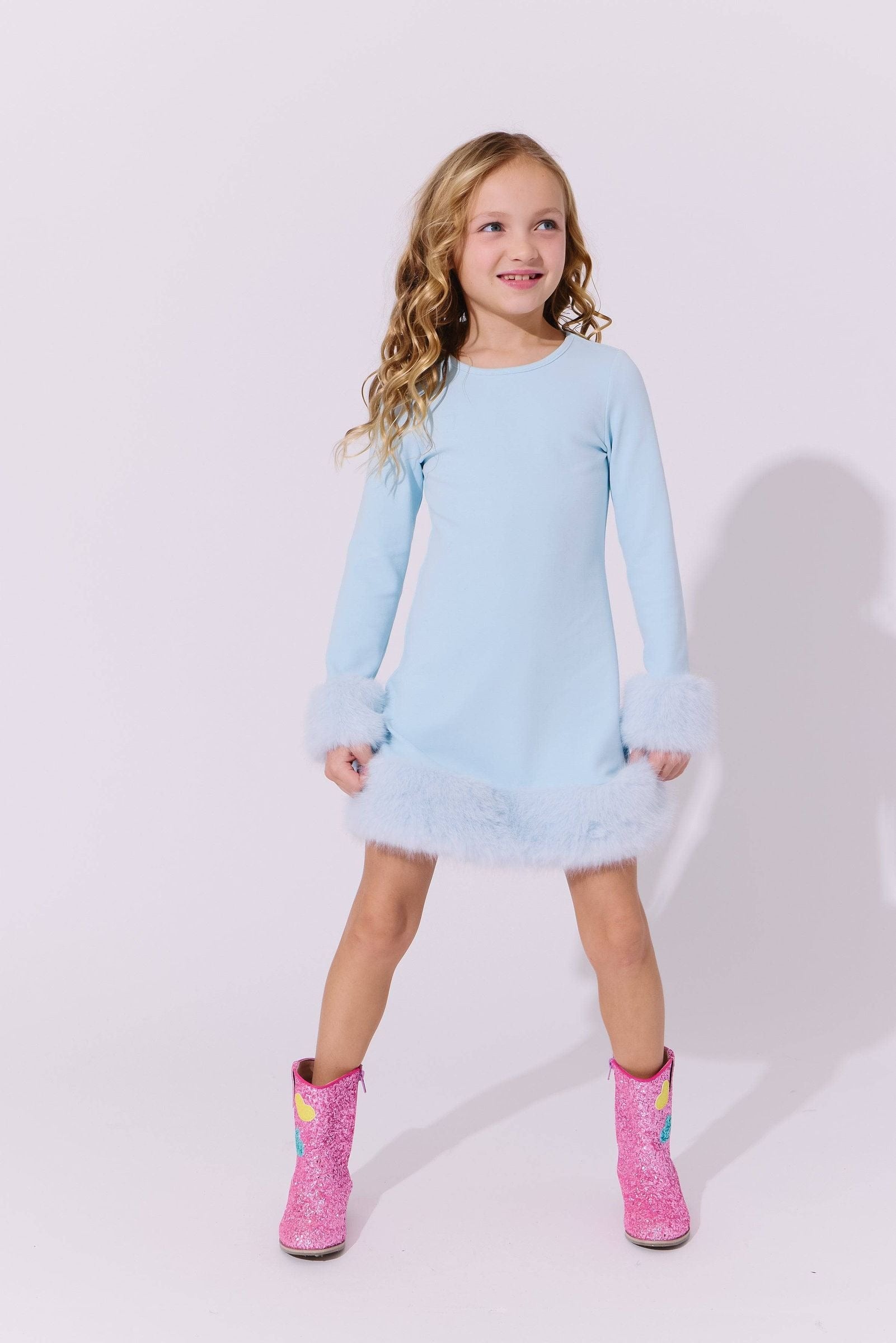 A young girl with blonde curly hair stands against a light gray studio background wearing a light blue long-sleeve dress with fluffy trim at the cuffs and hem, paired with pink glittery boots featuring rainbow accents.