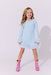 A young girl with blonde curly hair stands against a light gray studio background wearing a light blue long-sleeve dress with fluffy trim at the cuffs and hem, paired with pink glittery boots featuring rainbow accents.