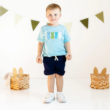 Child wearing a light blue t-shirt with colorful graphics and dark shorts, standing in a decorated room.