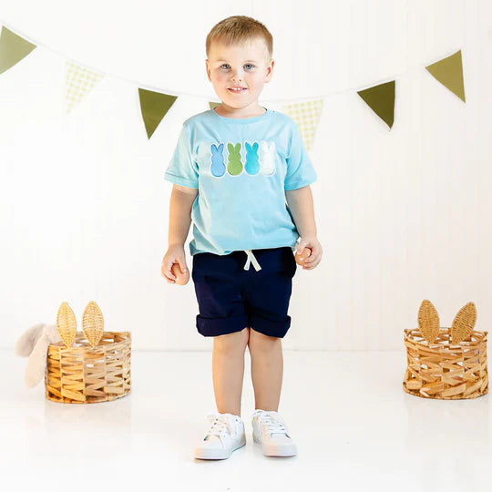Child wearing a light blue t-shirt with colorful graphics and dark shorts, standing in a decorated room.