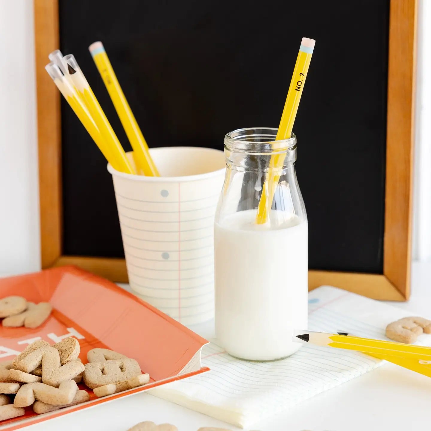 Glass bottle of milk with a yellow pencil, cup with more pencils, and cookies on a table.