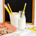 Glass bottle of milk with a yellow pencil, cup with more pencils, and cookies on a table.