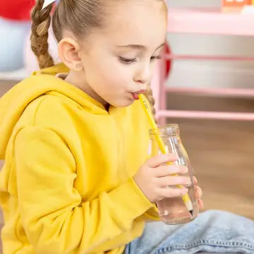 Child in a yellow hoodie drinking from a glass with a straw indoors.