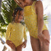 Two children in yellow swimsuits standing under a shower with palm trees in the background.