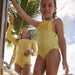 Two children in yellow swimsuits standing under a shower with palm trees in the background.