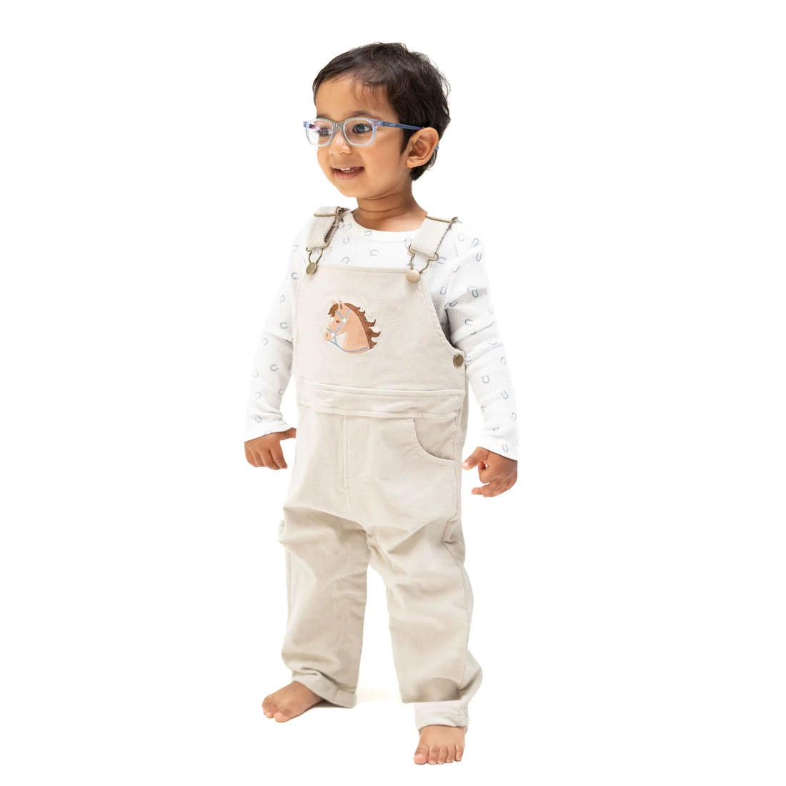 A toddler standing against a white background, wearing a light beige corduroy overall romper over a white long-sleeve shirt with a small hedgehog graphic on the chest.