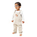 A toddler standing against a white background, wearing a light beige corduroy overall romper over a white long-sleeve shirt with a small hedgehog graphic on the chest.