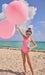 Child in a pink swimsuit holding pink balloons on a beach with blue water and sky.