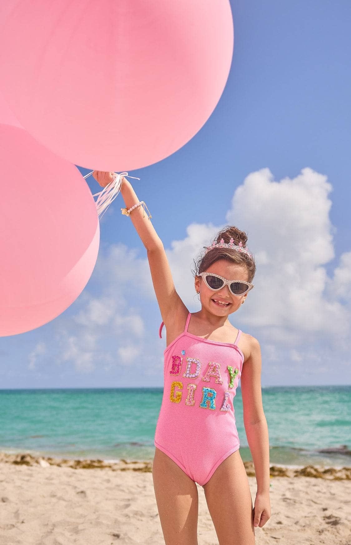 Child in a pink swimsuit with colorful letters on a beach holding pink balloons.
