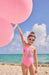Child in a pink swimsuit with colorful letters on a beach holding pink balloons.