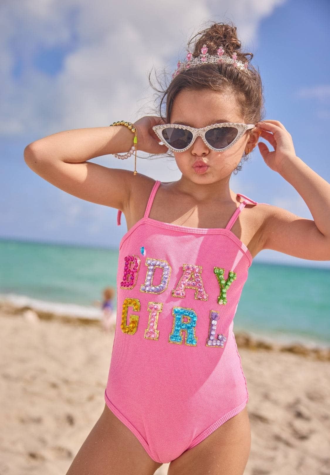 Young girl in a pink swimsuit with 'B-Day Girl' text on a beach
