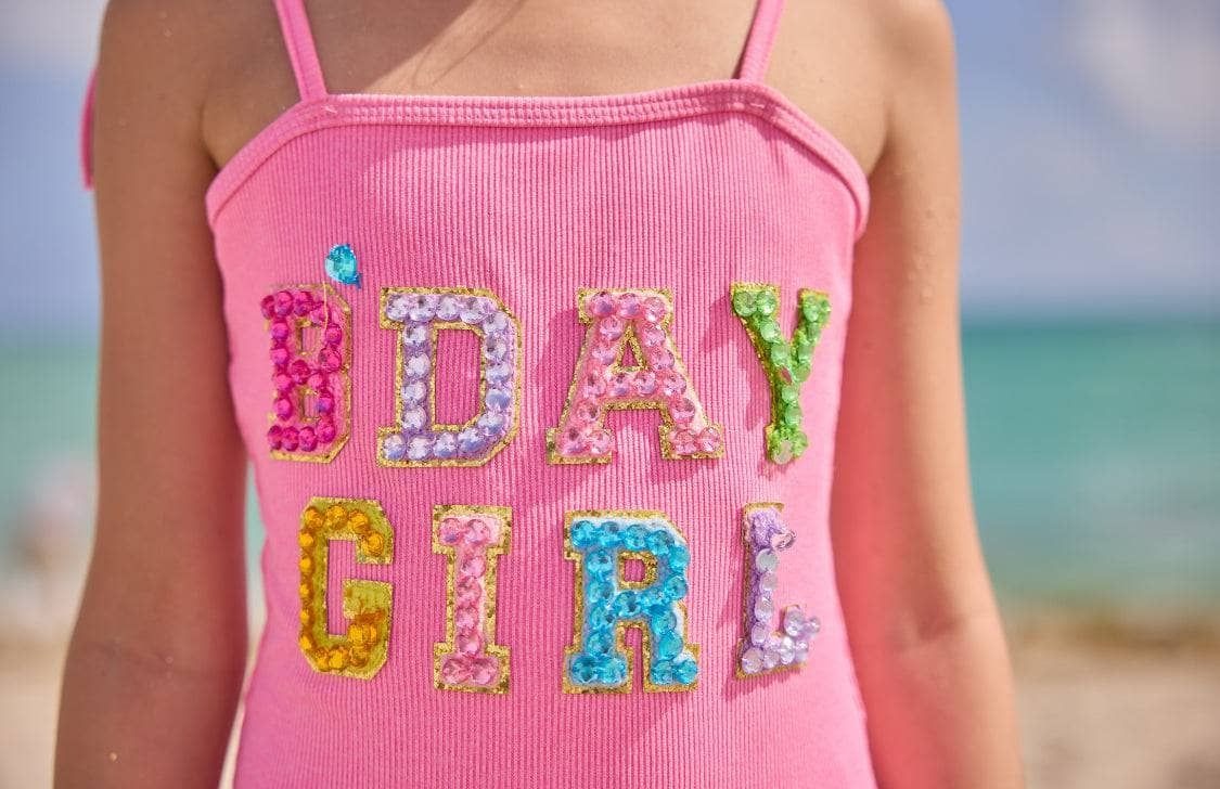 Pink tank top with 'B'DAY GIRL' text in colorful sequins on a beach.
