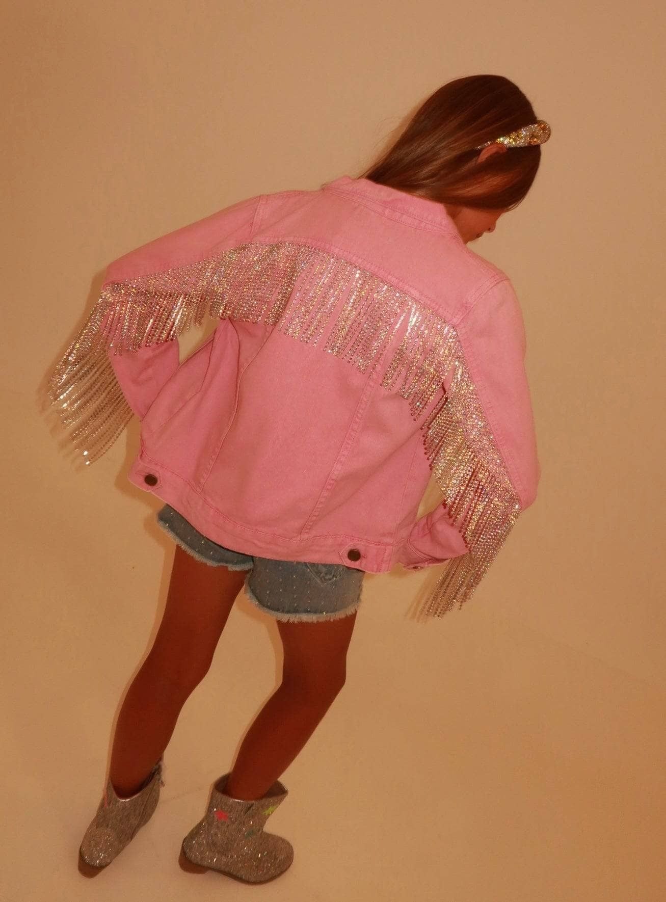 A child wearing a pink denim jacket with silver fringe across the shoulders and upper back, paired with denim shorts and glittery gray boots, photographed against a plain beige wall.
