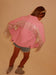 A child wearing a pink denim jacket with silver fringe across the shoulders and upper back, paired with denim shorts and glittery gray boots, photographed against a plain beige wall.