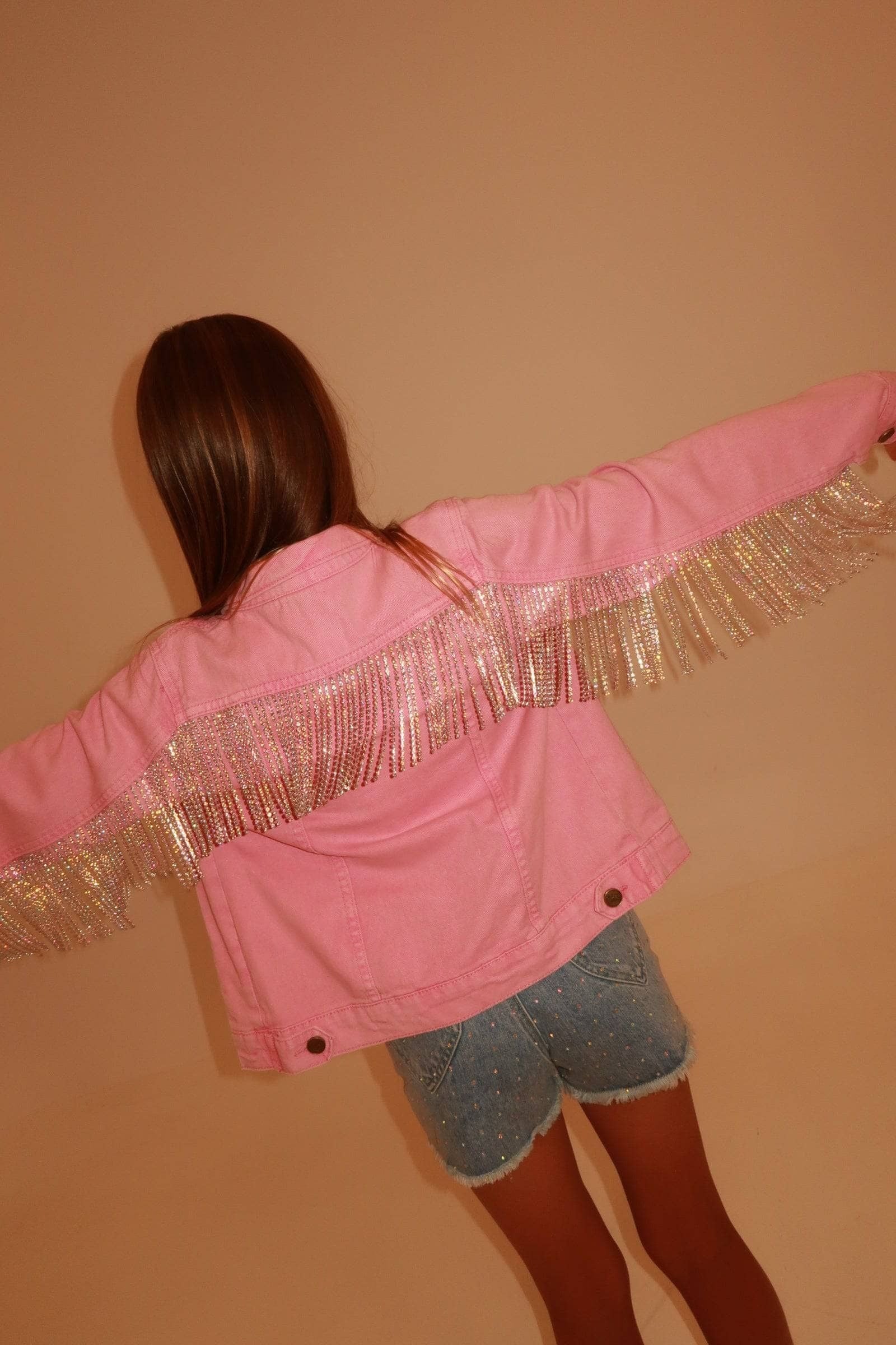 A person viewed from behind wearing a pink jacket with metallic fringe across the shoulders and upper back, paired with denim shorts, against a plain beige wall.