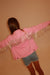 A person viewed from behind wearing a pink jacket with metallic fringe across the shoulders and upper back, paired with denim shorts, against a plain beige wall.