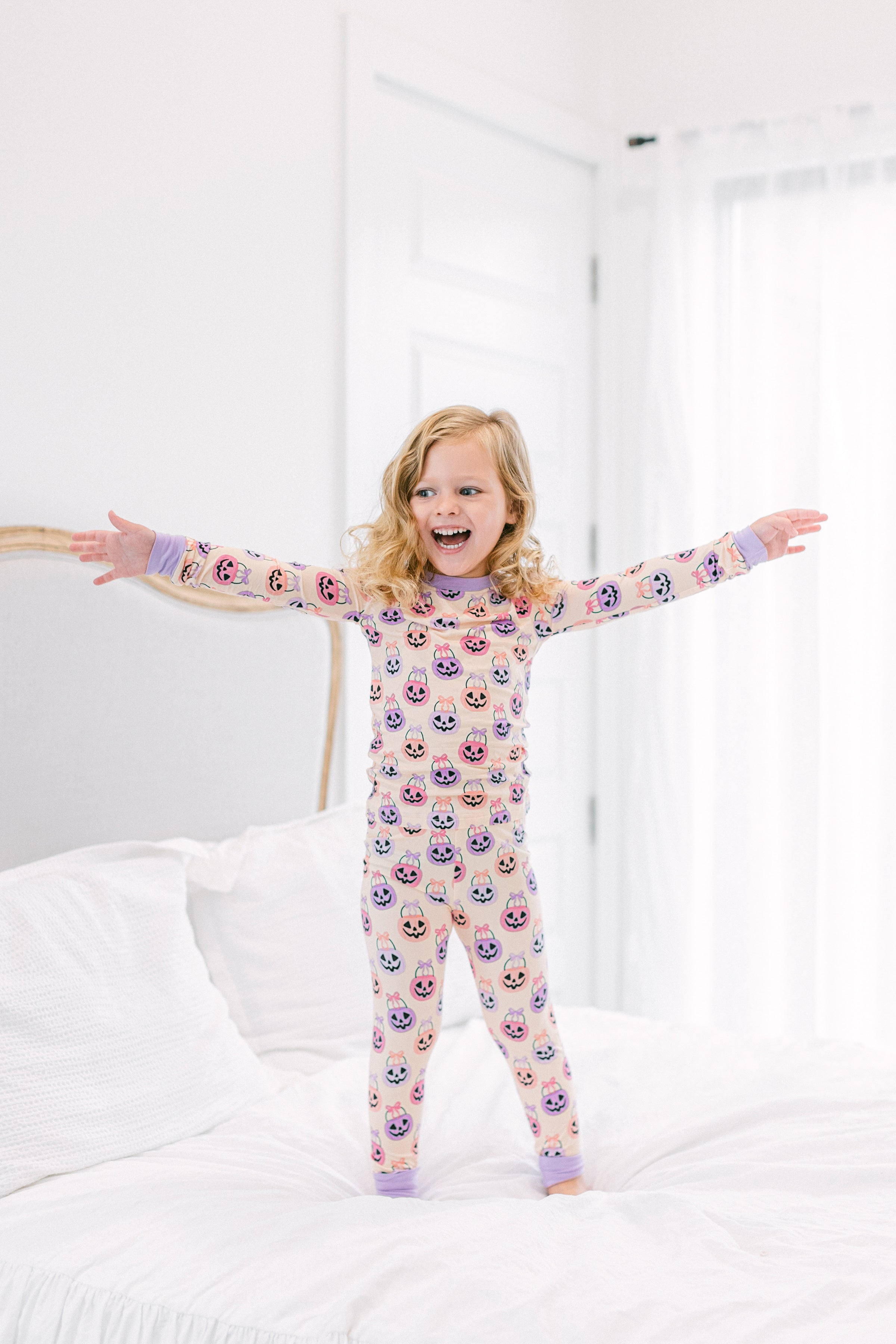 A young girl with blonde hair stands on a white bed with her arms outstretched, wearing a light pink long-sleeve pajama set covered i a colorful pattern of Halloween baskets.