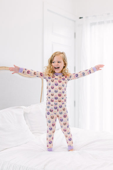 A young girl with blonde hair stands on a white bed with her arms outstretched, wearing a light pink long-sleeve pajama set covered i a colorful pattern of Halloween baskets.