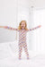 A young girl with blonde hair stands on a white bed with her arms outstretched, wearing a light pink long-sleeve pajama set covered i a colorful pattern of Halloween baskets.
