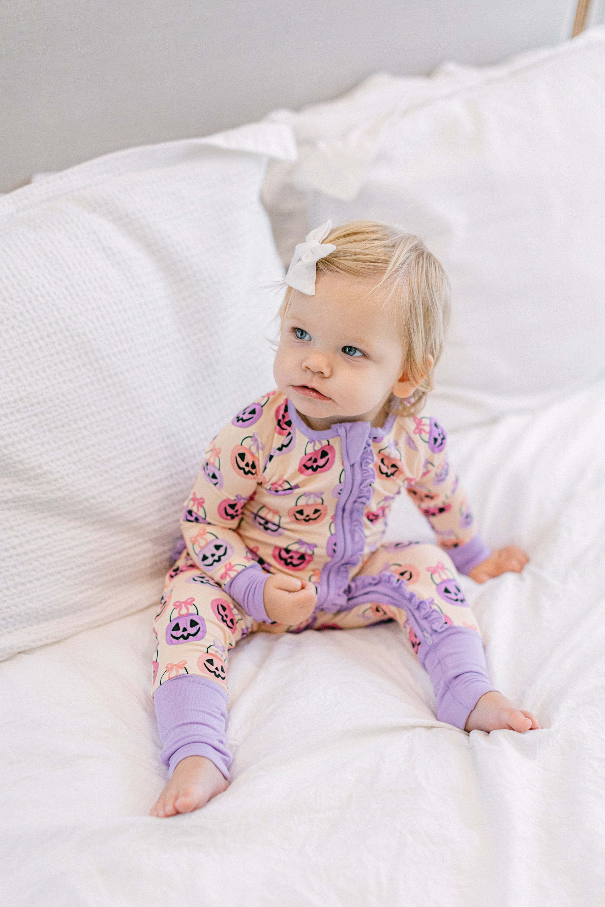 A toddler girl with blonde hair and a white bow sits on a white bed wearing a light pink and purple onesie pajama with pumpkin print