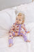A toddler girl with blonde hair and a white bow sits on a white bed wearing a light pink and purple onesie pajama with pumpkin print