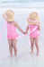 Two children in pink swimsuits and straw hats holding hands on a beach.