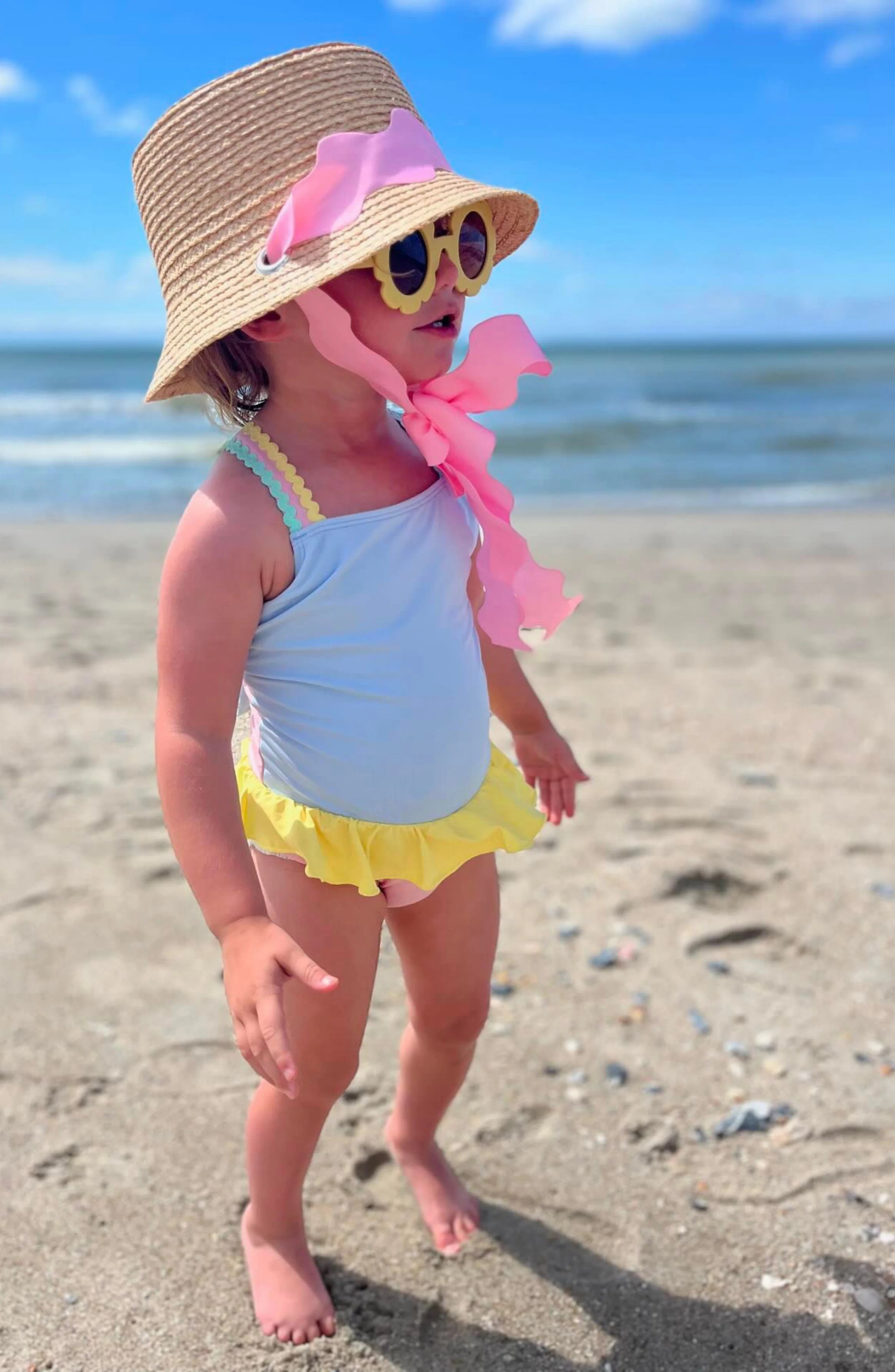 Child on a beach wearing a straw hat, sunglasses, and a swimsuit with pink accents.