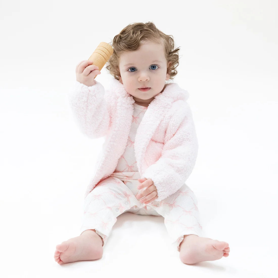 Baby wearing a soft pink chenille hoodie and patterned outfit while sitting barefoot on a white background. Angel Dear outerwear for cozy everyday wear.