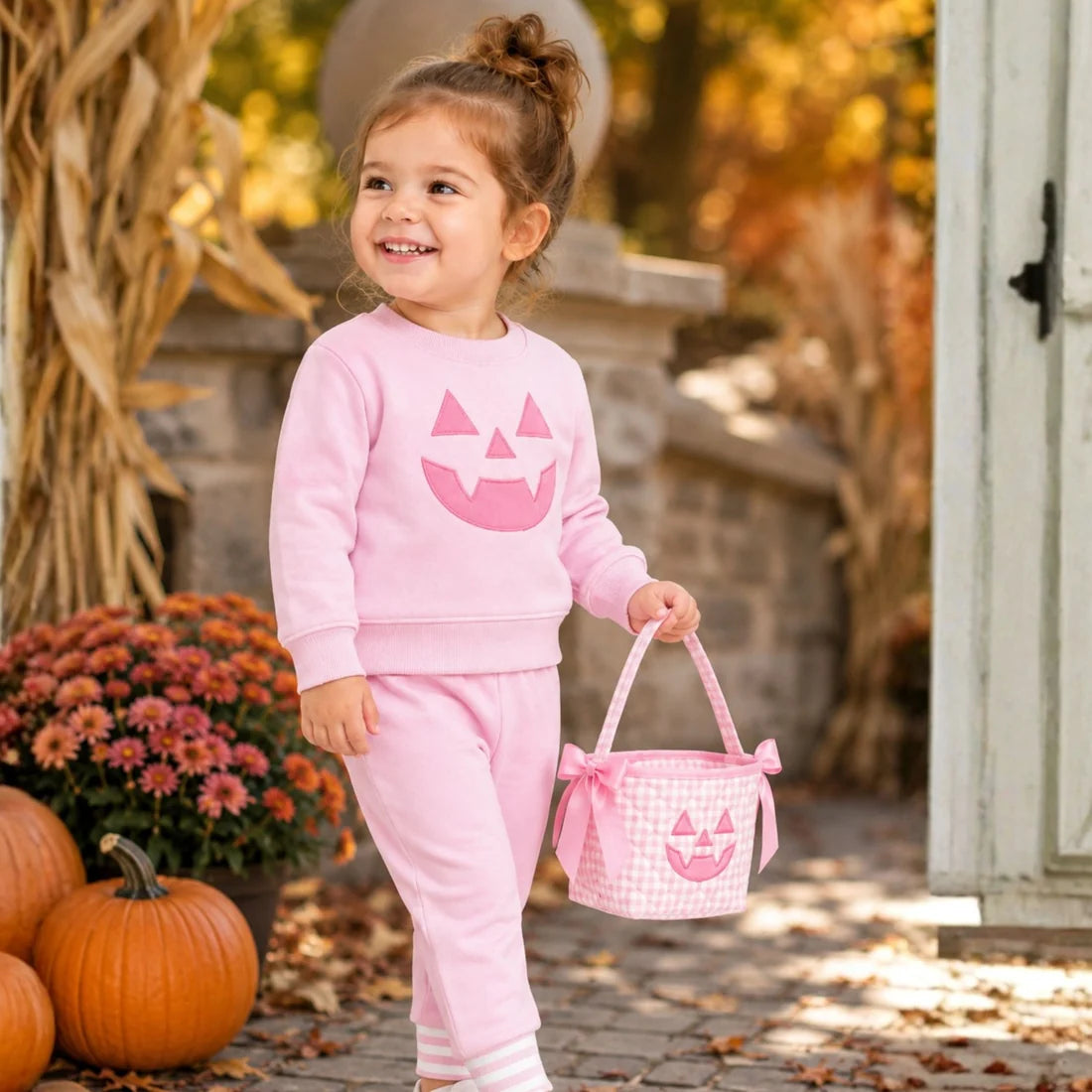 A young girl holding apink gingham quilted mini bucket bag with a pink satin handle, bow details on the sides, and an embroidered jack-o'-lantern face on the front.