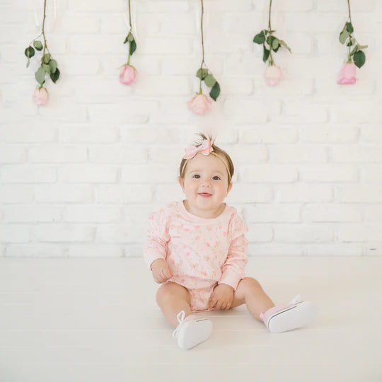 Baby sitting on a white floor with a white brick wall background, wearing a pink outfit and headband.