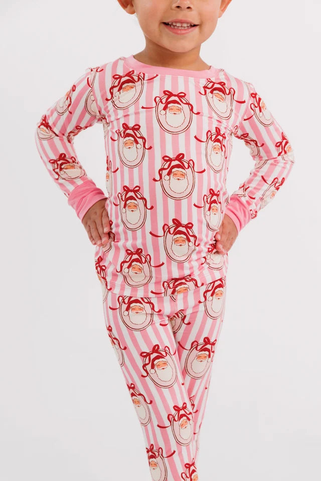 A child wearing a pink and white striped long-sleeve pajama set with a red Santa Claus face print, standing against a light gray studio background.