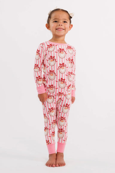 A young girl wearing a two-piece children's pajama set featuring a pink and white striped pattern with santa faces wearing red bows, complete with solid pink cuffs and collar.
