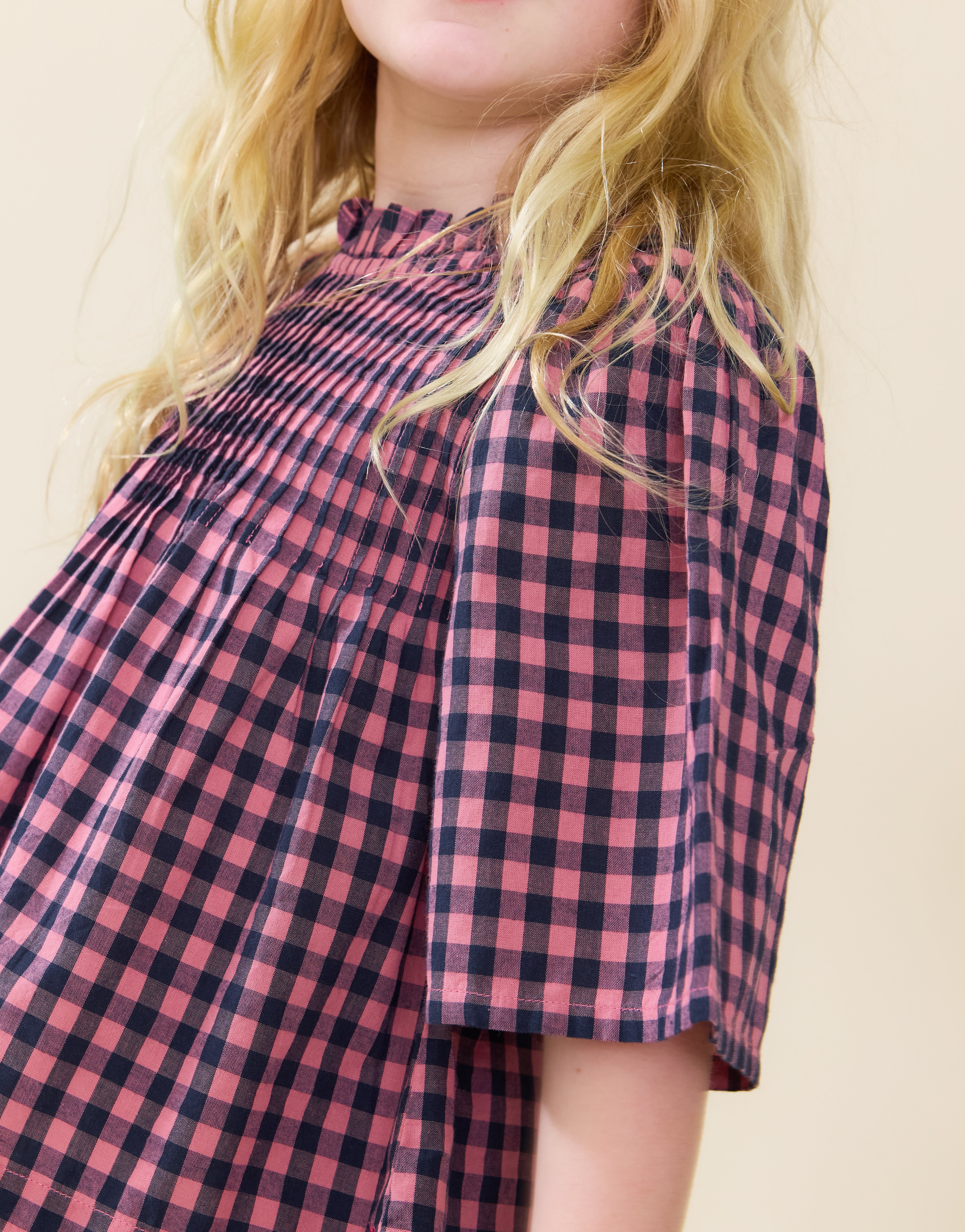 A close-up of a child wearing a pink and navy blue gingham check top with a smocked neckline and short puff sleeves, set against a plain beige background.