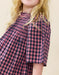 A close-up of a child wearing a pink and navy blue gingham check top with a smocked neckline and short puff sleeves, set against a plain beige background.