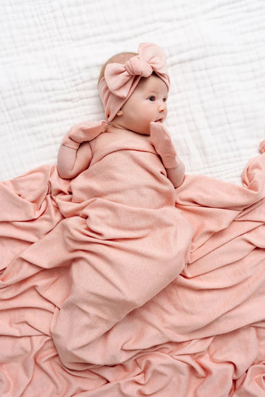 A baby featuring a square blanket, a matching bow headband, and a pair of mittens, all in a soft pink color, displayed on a solid white background.