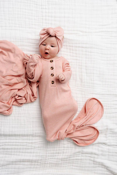 A newborn baby wearing a dusty pink button-front bodysuit and matching bow headband, lying on a white textured muslin blanket with a matching pink blanket beside them.