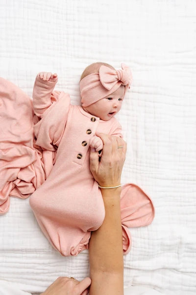 A baby wearing a dusty pink button-front bodysuit and matching bow headband, being held on a white textured blanket by an adult's hands.