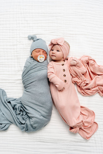 Two newborn babies lying side by side on a white textured blanket. The baby on the left is swaddled in a dusty blue blanket and wearing a matching blue bonnet with a pacifier.