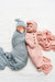 Two newborn babies lying side by side on a white textured blanket. The baby on the left is swaddled in a dusty blue blanket and wearing a matching blue bonnet with a pacifier.