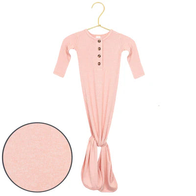 A baby sleep sack in light pink with a small button placket, hanging on a gold hanger against a white background, with a circular fabric texture inset in the lower left corner.