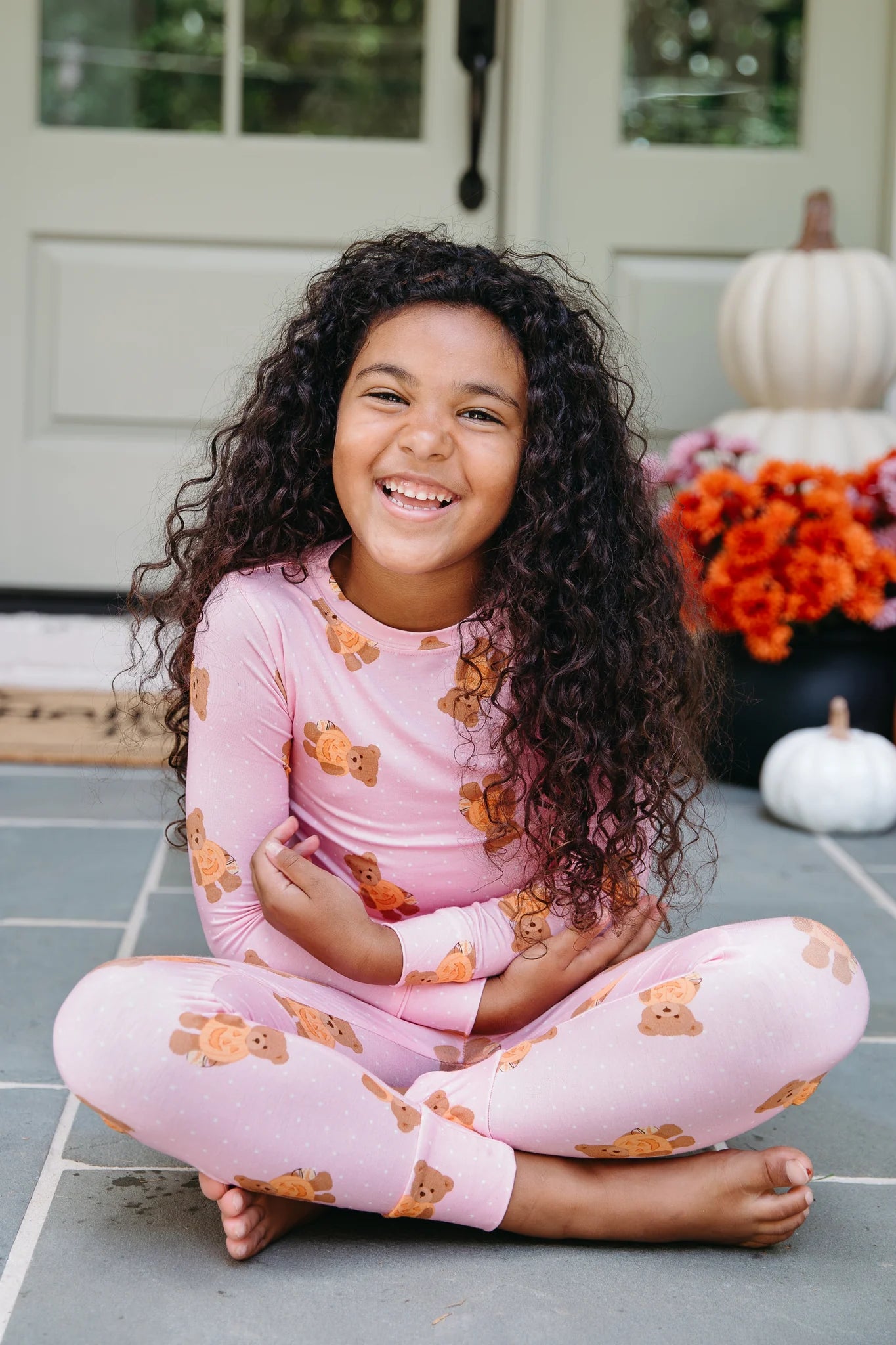 A young girl with long curly dark hair sits cross-legged on a gray tiled porch wearing a light pink long-sleeve pajama top and matching pants, both printed with small teddy bear motifs. A white door, pumpkins, and orange flowers are blurred in the background.