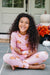 A young girl with long curly dark hair sits cross-legged on a gray tiled porch wearing a light pink long-sleeve pajama top and matching pants, both printed with small teddy bear motifs. A white door, pumpkins, and orange flowers are blurred in the background.