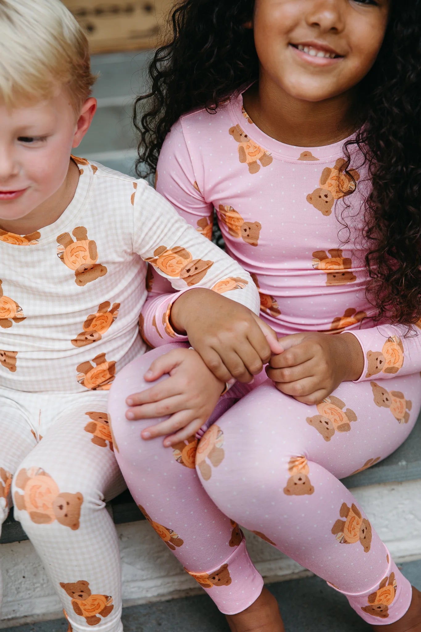 Two children wearing matching long-sleeve pajamas with a teddy bear print, sitting together on outdoor steps. The child on the left wears a white pajama set, and the child on the right wears a light pink pajama set.